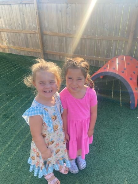 Two young girls stand smiling on artificial grass in a sunny outdoor play area. One wears a patterned dress with blue trim, the other a pink dress. A small red playground structure with black dots is behind them.