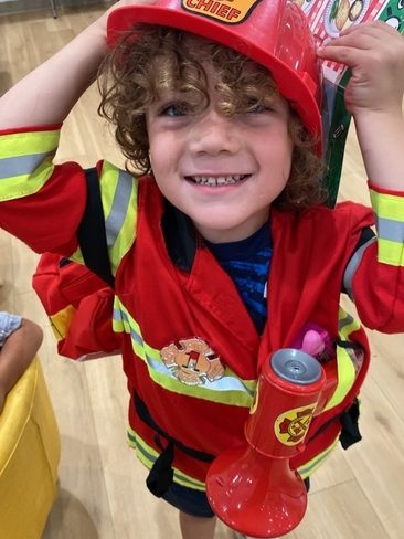 A smiling young child with curly hair wears a red firefighter costume and helmet labeled "FIRE CHIEF." The child holds the helmet and carries a toy megaphone, standing indoors on a light wood floor.