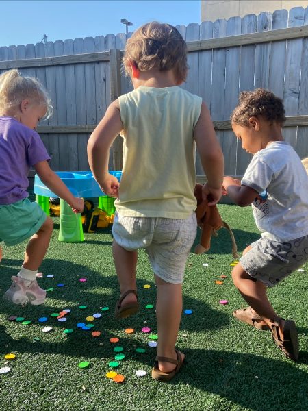 Four young children play outside on artificial grass, picking up or stepping on colorful round tokens scattered on the ground, with wooden fencing and a small play structure in the background.