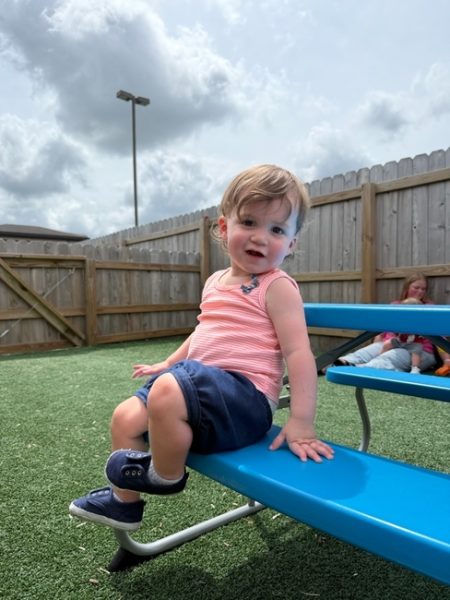 A young child in a pink striped tank top and navy shorts sits on a bright blue bench outside on artificial grass, with a wooden fence and cloudy sky in the background. Another child sits on the ground nearby.