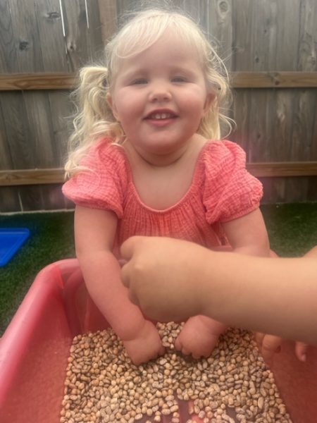 A smiling young girl with blonde hair and a pink dress has her hands in a bin filled with dry beans, while another child's hand reaches in. They are outdoors near a wooden fence.