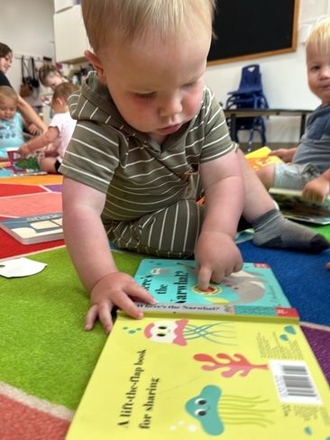 A baby in a striped outfit sits on a colorful rug, focused on exploring board books with sea creature illustrations. Other young children and toys are visible in the background.