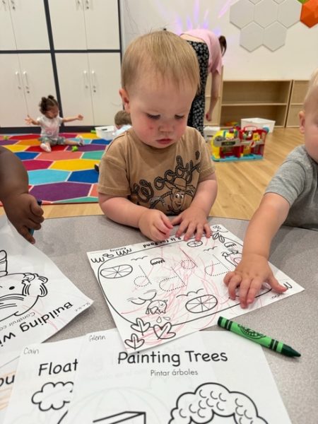 A toddler sits at a table coloring on a worksheet with red crayon. Another child is drawing beside him. In the background, a child plays on a colorful rug and toys are scattered around the room.
