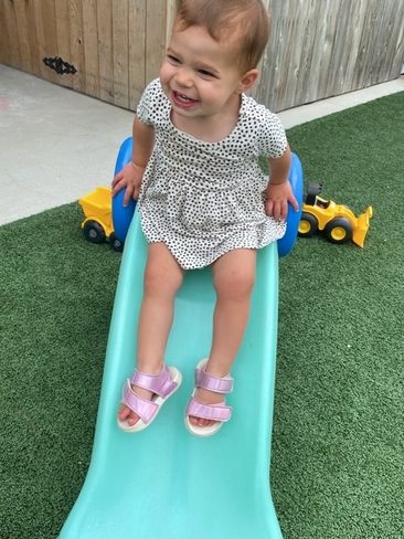 A smiling toddler in a white dress with black polka dots sits at the top of a small blue and green slide, with toy construction trucks on artificial grass in the background.
