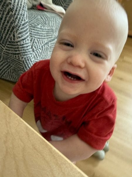 A smiling baby with light skin and short blond hair, wearing a red shirt, stands near a wooden surface with a patterned chair in the background.