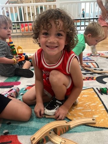 A young child with curly hair, wearing a red sleeveless shirt and shorts, smiles at the camera while kneeling on a colorful play rug in a classroom. Other children play with wooden toys in the background.
