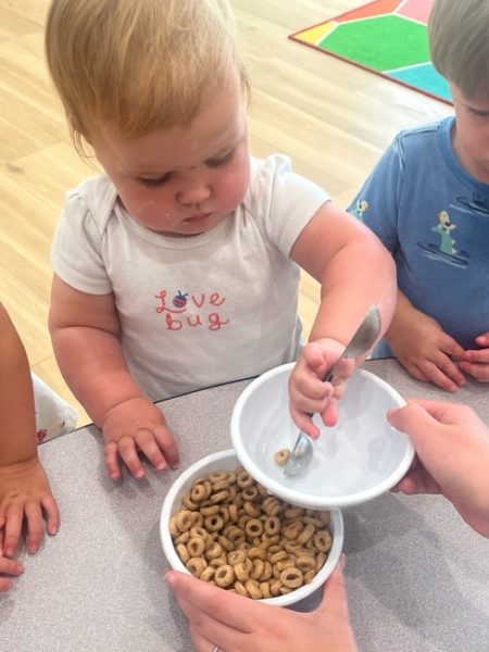 A toddler scoops cereal from a bowl into her own bowl with a spoon, while standing at a table with other children. She wears a white "Love bug" shirt and looks focused on the task.