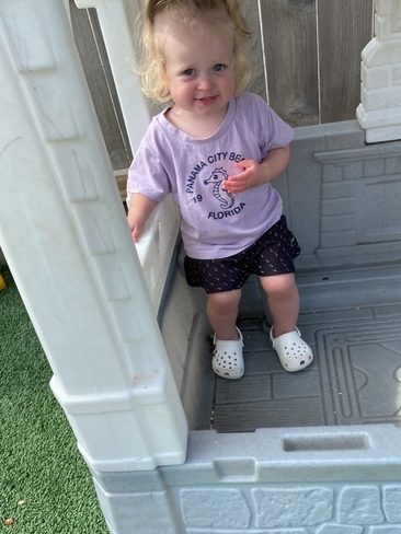 A toddler with blonde hair in pigtails stands inside a plastic playhouse. She is smiling, wearing a purple t-shirt, black shorts with white dots, and white clogs. The ground outside is green artificial turf.