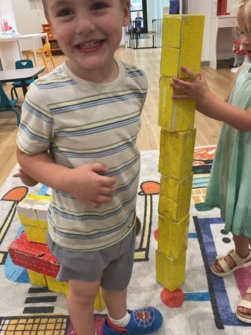 A young boy smiles at the camera while standing next to a tall tower made of yellow toy blocks. Another child’s hands are visible stacking the blocks. They are in a brightly lit playroom with colorful furniture and a patterned rug.