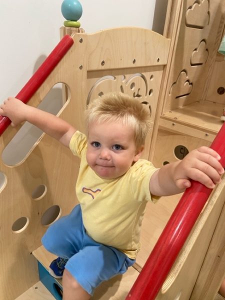 A young child with blond hair, wearing a yellow shirt and blue shorts, holds onto red railings while sitting on wooden playground equipment indoors.