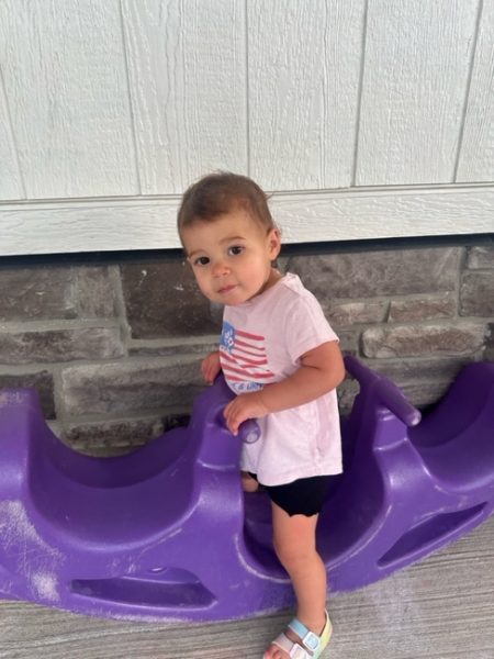 A toddler wearing a light pink shirt with an American flag print, dark shorts, and sandals stands next to a purple plastic rocker in front of a stone and wood wall.