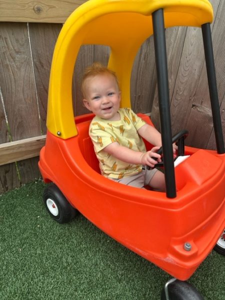 A smiling toddler with light hair sits inside a red and yellow toy car on artificial grass, next to a wooden fence, holding the steering wheel.