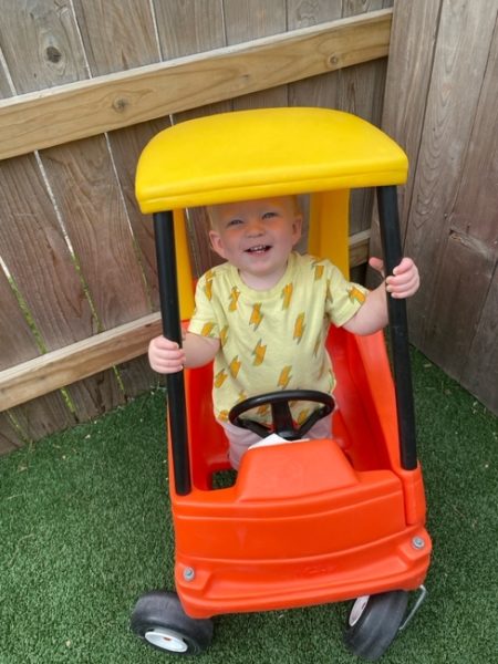 A smiling toddler wearing a yellow shirt with lightning bolts stands inside a red and yellow toy car on green artificial grass, next to a wooden fence.