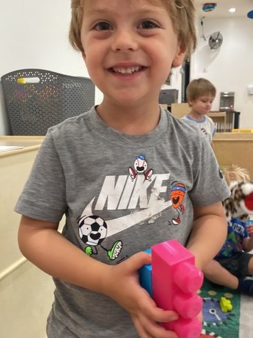 A young child with light brown hair, wearing a grey Nike t-shirt, smiles at the camera while holding colorful building blocks in a playroom setting. Another child is visible in the background.
