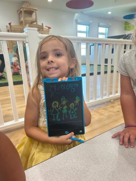 A young girl in a yellow dress smiles while holding up a drawing tablet displaying colorful stick figure drawings of people and a house. She is sitting at a table in a bright indoor play area.
