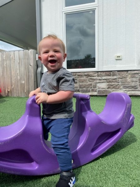 A smiling toddler stands on a purple plastic seesaw outdoors on artificial grass, with a wooden fence and a white building in the background.