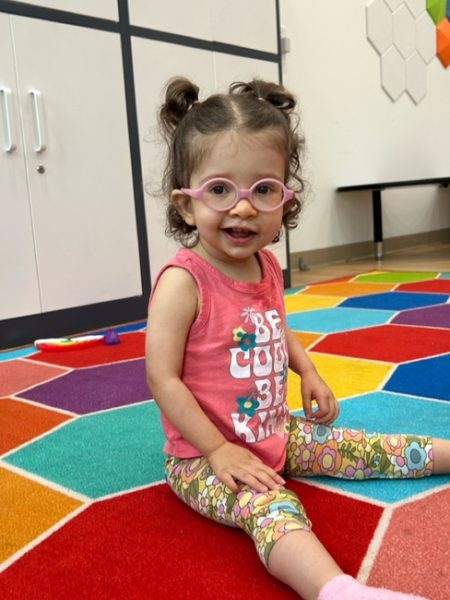 A young girl with curly hair and pink glasses sits smiling on a colorful hexagon-patterned carpet. She wears a pink shirt that says "Be Cool Be Kind" and floral leggings. Shelves and wall tiles are in the background.