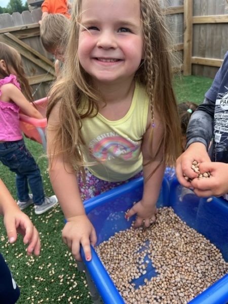 A young girl smiles while playing with dry beans in a blue plastic bin outdoors, surrounded by other children.