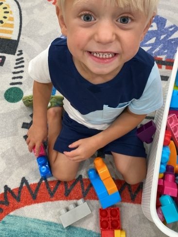 A young boy with blonde hair and blue eyes smiles up at the camera while sitting on a colorful carpet, playing with large, colorful plastic building blocks.
