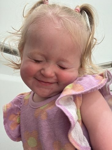 A young child with blonde hair in pigtails smiles with eyes closed, wearing a purple top with yellow and pink flowers. The background is plain and light-colored.