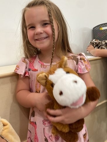 A smiling young girl with long, partially braided hair wears a pink dress with horse prints and holds a brown and white stuffed horse toy. She stands indoors near a wooden surface with toys in the background.