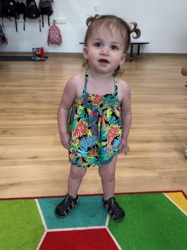 A toddler with pigtails, wearing a colorful floral romper and black sandals, stands on a brightly colored rug in a room with a wooden floor and backpacks hanging on the wall behind.