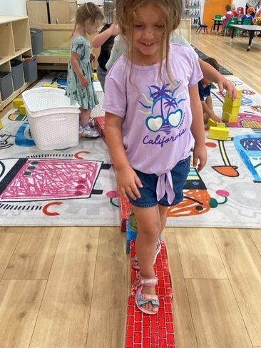 A young girl in a "California" t-shirt walks carefully on a path of red toy blocks in a colorful classroom, with other children playing in the background.