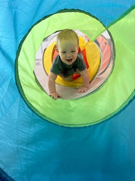 A smiling baby crawls through a colorful play tunnel, looking out through a circular opening in the blue and green fabric toward the camera.