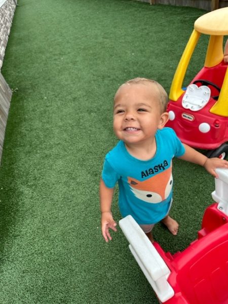 A smiling toddler wearing a blue shirt with a fox design stands barefoot on green artificial grass, holding onto a red and white toy car. Another red and yellow toy car is in the background.