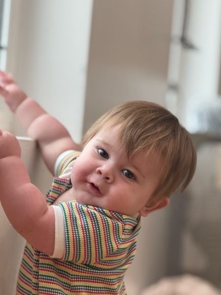A baby with light brown hair, wearing a colorful striped shirt, stands and holds onto a surface while looking at the camera with a slight smile. The background is softly blurred.