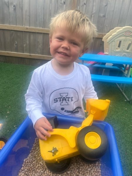 A young boy smiling while playing with a yellow toy truck in a blue bin filled with beans, outdoors on green artificial grass with a wooden fence and playground equipment in the background.