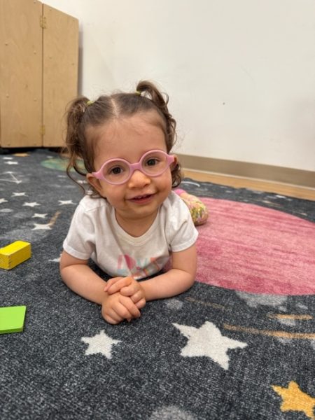 A young child with brown hair in pigtails, wearing pink glasses and a white shirt, smiles while lying on a space-themed carpet with star and planet designs. Colorful toy blocks are nearby.