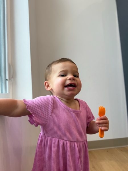 A toddler in a pink dress stands by a window, smiling and holding an orange plastic fork in one hand. The background features light-colored walls and a wooden floor.