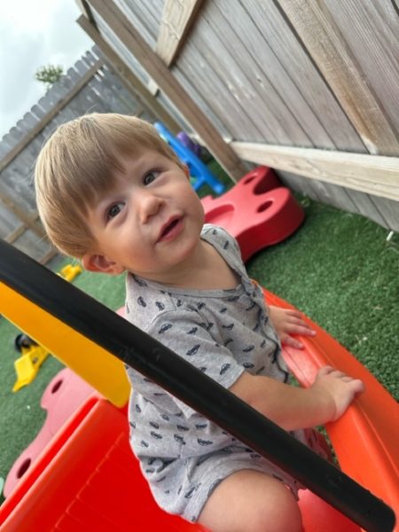 A smiling toddler with light brown hair sits inside a red and yellow toy car on artificial grass in a fenced backyard, looking up toward the camera.