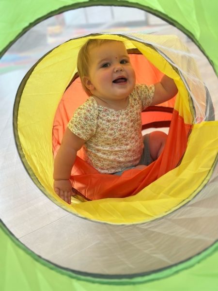 A smiling baby sits inside a colorful play tunnel, looking up through the circular opening. The tunnel is bright yellow, orange, and green, and the baby wears a floral-patterned shirt.