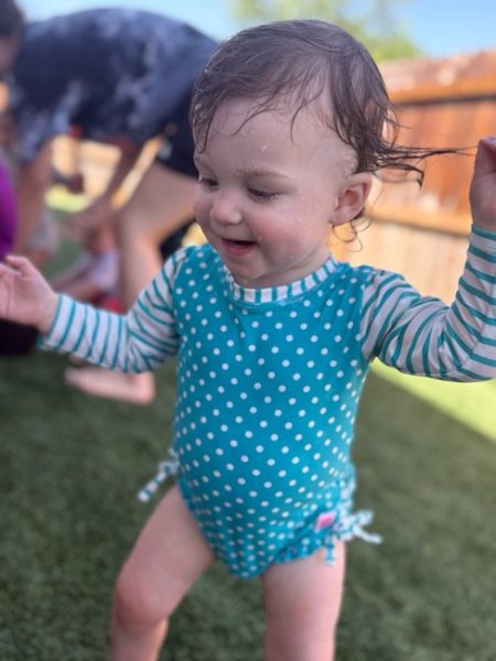 A toddler in a blue and white polka-dot swimsuit smiles while playing outside on grass. The child’s hair is wet, and other people are blurred in the background near a wooden fence.