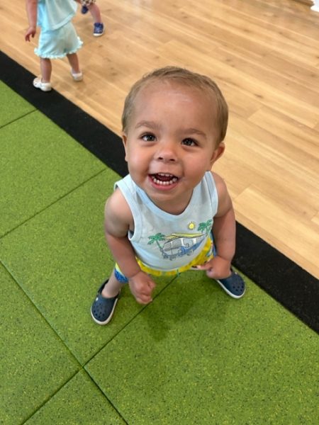 A young toddler smiles up at the camera while standing on a green play mat. He wears a tank top, yellow shorts, and blue shoes. Another child is visible in the background on a wooden floor.