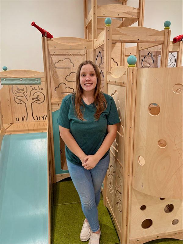 A smiling girl with long brown hair in a green shirt and blue jeans stands in front of a wooden indoor playground structure with a slide and decorative cut-outs.