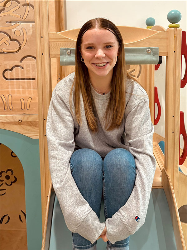 A teenage girl with straight brown hair, wearing a gray sweatshirt and blue jeans, sits smiling at the bottom of an indoor wooden slide in a play area decorated with carved patterns.