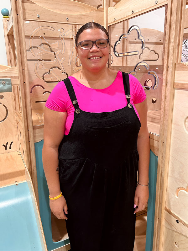 A woman with glasses and tied-back hair, wearing a bright pink shirt under black overalls, stands smiling in front of a light wooden play structure with cloud and tree designs.